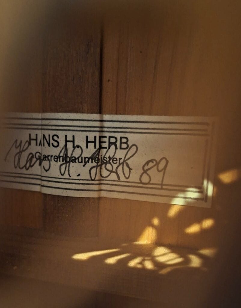 Eine Nahaufnahme des Etiketts auf der Hans Herb 1989 Vihuela zeigt den Schriftzug „HANS H. HERB, Glasrestaurierungsmeister“ mit einer handschriftlichen Unterschrift und der Zahl 89, während warmes Licht und Schatten die Sicht teilweise verdecken. Eine Nahaufnahme des Etiketts auf der Hans Herb 1989 Vihuela zeigt den Schriftzug „HANS H. HERB, Glasrestaurierungsmeister“ mit einer handschriftlichen Unterschrift und der Zahl 89, während warmes Licht und Schatten die Sicht teilweise verdecken.