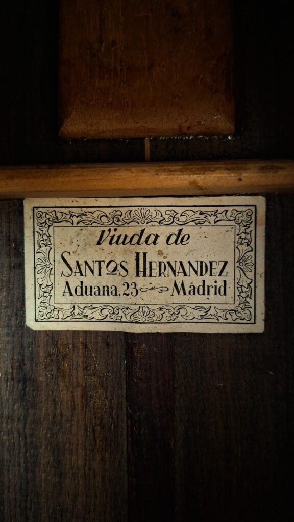 Ein Vintage-Etikett auf der Holzoberfläche der „Angel Alonso 1936 Klassische Gitarre – Santos Hernandez“ trägt die Aufschrift „Viuda de Santos Hernandez, Aduana, 23, Madrid“, umgeben von einer dekorativen Blumenbordüre. Ein Vintage-Etikett auf der Holzoberfläche der „Angel Alonso 1936 Klassische Gitarre – Santos Hernandez“ trägt die Aufschrift „Viuda de Santos Hernandez, Aduana, 23, Madrid“, umgeben von einer dekorativen Blumenbordüre.