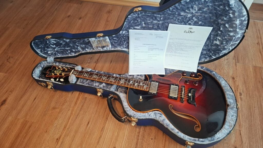 Eine DeLaHaye-Elektroakustikgitarre aus dem Jahr 1995 mit Sunburst-Lackierung liegt in einem offenen, mit Samt ausgekleideten Hartschalenkoffer auf einem Holzboden, wobei zwei Dokumente quer über den Korpus der Gitarre gelegt sind. Eine DeLaHaye-Elektroakustikgitarre aus dem Jahr 1995 mit Sunburst-Lackierung liegt in einem offenen, mit Samt ausgekleideten Hartschalenkoffer auf einem Holzboden, wobei zwei Dokumente quer über den Korpus der Gitarre gelegt sind.
