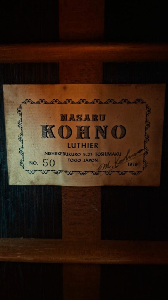 Auf einer Nahaufnahme eines Gitarrenetiketts ist zu lesen: „Masaru Kohno, Gitarrenbauer, Nishikebukuro 5-27, Toshima-ku, Tokio, Japan, Modell 50, 1979“, mit einer Unterschrift auf der rechten Seite. Das Etikett ist auf der dunklen Holzoberfläche einer klassischen Gitarre vom Typ „Masaru Kohno Modell 50, 1979“ angebracht. Auf einer Nahaufnahme eines Gitarrenetiketts ist zu lesen: „Masaru Kohno, Gitarrenbauer, Nishikebukuro 5-27, Toshima-ku, Tokio, Japan, Modell 50, 1979“, mit einer Unterschrift auf der rechten Seite. Das Etikett ist auf der dunklen Holzoberfläche einer klassischen Gitarre vom Typ „Masaru Kohno Modell 50, 1979“ angebracht.