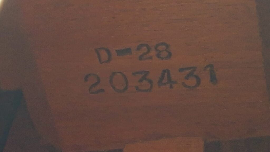 Nahaufnahme der Holzoberfläche einer Martin D-28 Akustikgitarre aus dem Jahr 1965, auf der der schwarze Schriftzug „D-28 203431“ eingeprägt oder aufgedruckt ist. Die Oberfläche wirkt leicht rau und wird von warmem Licht beleuchtet. Nahaufnahme der Holzoberfläche einer Martin D-28 Akustikgitarre aus dem Jahr 1965, auf der der schwarze Schriftzug „D-28 203431“ eingeprägt oder aufgedruckt ist. Die Oberfläche wirkt leicht rau und wird von warmem Licht beleuchtet.