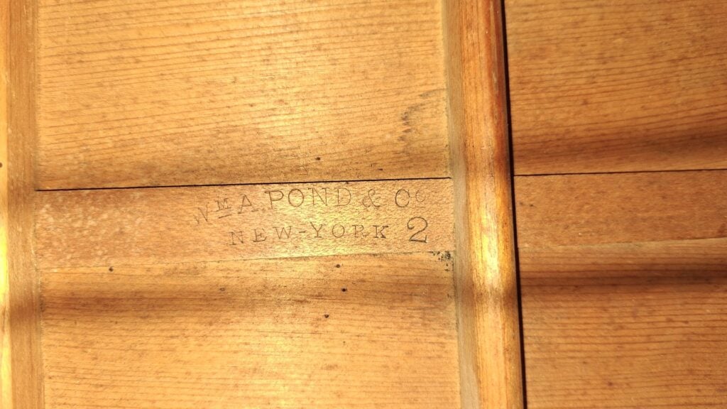 Nahaufnahme der Holzoberfläche einer James-Ashborn-Parlor-Akustikgitarre aus dem Jahr 1850, auf der zwischen zwei vertikalen Holzlatten der Stempel „J.A. Pond & Co. New York 2“ in verblasster schwarzer Tinte zu sehen ist. Nahaufnahme der Holzoberfläche einer James-Ashborn-Parlor-Akustikgitarre aus dem Jahr 1850, auf der zwischen zwei vertikalen Holzlatten der Stempel „J.A. Pond & Co. New York 2“ in verblasster schwarzer Tinte zu sehen ist.