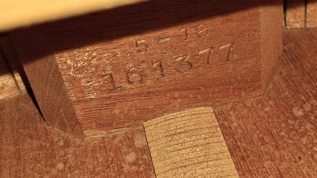 Nahaufnahme einer Holzoberfläche, auf der die Zahl 161377 sowie teilweise erkennbare Zahlen und Buchstaben eingraviert sind, möglicherweise im Inneren einer Martin 5-18 Akustikgitarre aus dem Jahr 1958. Nahaufnahme einer Holzoberfläche, auf der die Zahl 161377 sowie teilweise erkennbare Zahlen und Buchstaben eingraviert sind, möglicherweise im Inneren einer Martin 5-18 Akustikgitarre aus dem Jahr 1958.