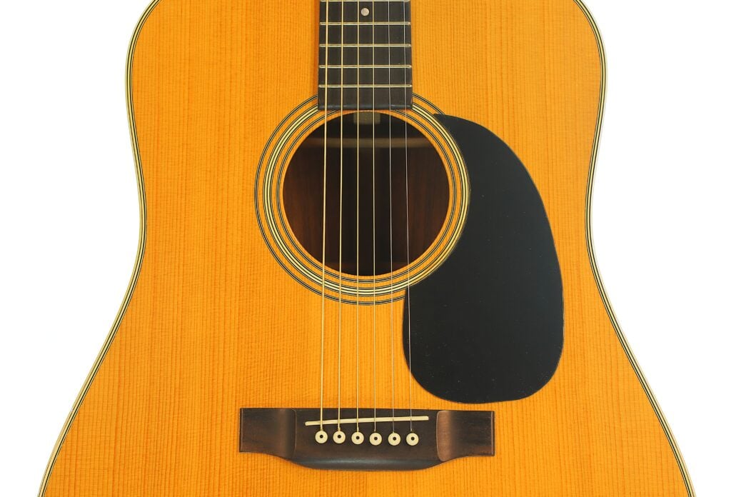 Nahaufnahme des Korpus einer Martin D-28 Akustikgitarre aus dem Jahr 1975 mit einem Steg aus Holz, einem runden Schallloch und einem schwarzen Schlagbrett. Nahaufnahme des Korpus einer Martin D-28 Akustikgitarre aus dem Jahr 1975 mit einem Steg aus Holz, einem runden Schallloch und einem schwarzen Schlagbrett.