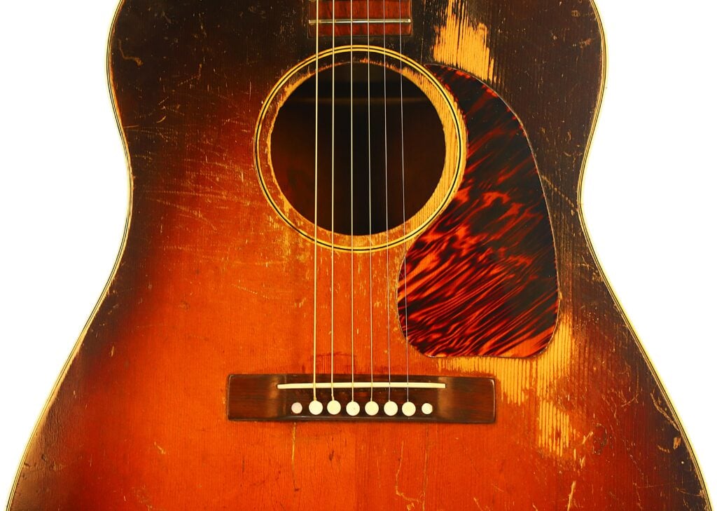 Nahaufnahme des Korpus einer Gibson J-45 Banner Akustikgitarre aus dem Jahr 1943 mit deutlichen Gebrauchsspuren, einer Sunburst-Lackierung, einem Pickguard in Schildpattoptik und sechs Stahlsaiten. Nahaufnahme des Korpus einer Gibson J-45 Banner Akustikgitarre aus dem Jahr 1943 mit deutlichen Gebrauchsspuren, einer Sunburst-Lackierung, einem Pickguard in Schildpattoptik und sechs Stahlsaiten.