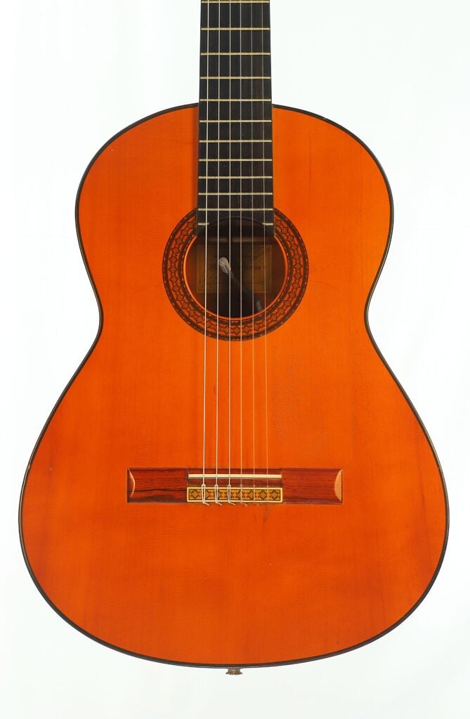 Eine Manuel Contreras 1a Flamenco-Gitarre aus dem Jahr 1977 mit einem hellorangefarbenen Korpus, einem dunklen Griffbrett und einer aufwendig gearbeiteten kreisförmigen Rosette um das Schallloch. Eine Manuel Contreras 1a Flamenco-Gitarre aus dem Jahr 1977 mit einem hellorangefarbenen Korpus, einem dunklen Griffbrett und einer aufwendig gearbeiteten kreisförmigen Rosette um das Schallloch.