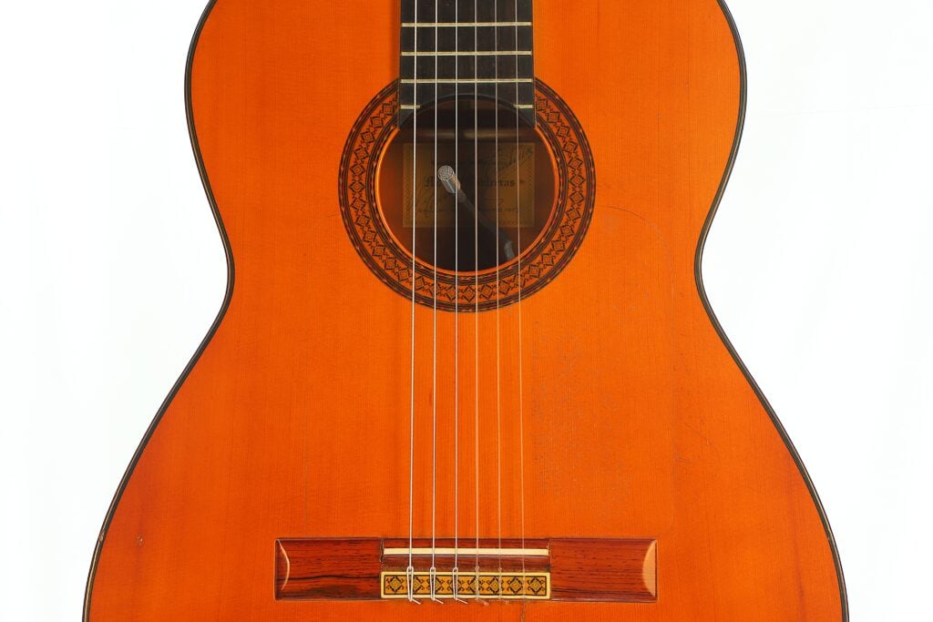 Nahaufnahme des Korpus einer Manuel Contreras 1a Flamenco-Gitarre aus dem Jahr 1977 mit einer dunklen, runden Rosette um das Schallloch und einem Holzsteg mit sechs Saiten. Nahaufnahme des Korpus einer Manuel Contreras 1a Flamenco-Gitarre aus dem Jahr 1977 mit einer dunklen, runden Rosette um das Schallloch und einem Holzsteg mit sechs Saiten.