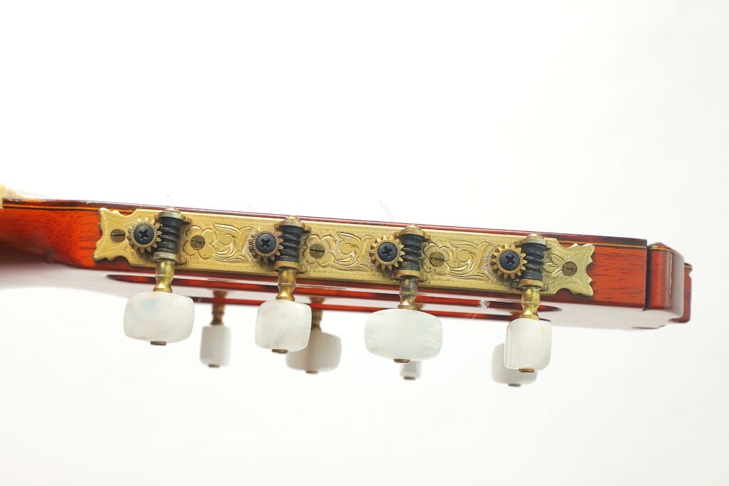 Kopfplatte einer 8-saitigen klassischen Gitarre von Jose Ramirez aus dem Jahr 1984 mit kunstvoll verzierten, goldfarbenen Stimmmechaniken und perlmuttfarbenen Stimmknöpfen. Kopfplatte einer 8-saitigen klassischen Gitarre von Jose Ramirez aus dem Jahr 1984 mit kunstvoll verzierten, goldfarbenen Stimmmechaniken und perlmuttfarbenen Stimmknöpfen.