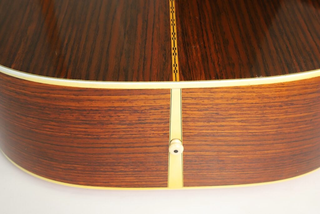 Nahaufnahme der Unterkante einer Martin D-28 Akustikgitarre aus dem Jahr 1975, die die Holzmaserung, die Einlage an der Mittelnaht und den Gurtstift zeigt. Nahaufnahme der Unterkante einer Martin D-28 Akustikgitarre aus dem Jahr 1975, die die Holzmaserung, die Einlage an der Mittelnaht und den Gurtstift zeigt.