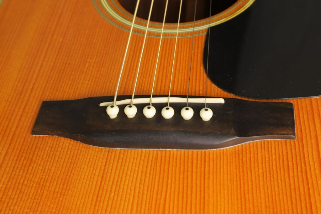 Nahaufnahme des Stegs einer Martin D-28 Akustikgitarre aus dem Jahr 1975 mit sechs Saiten und weißen Stegstiften. Nahaufnahme des Stegs einer Martin D-28 Akustikgitarre aus dem Jahr 1975 mit sechs Saiten und weißen Stegstiften.