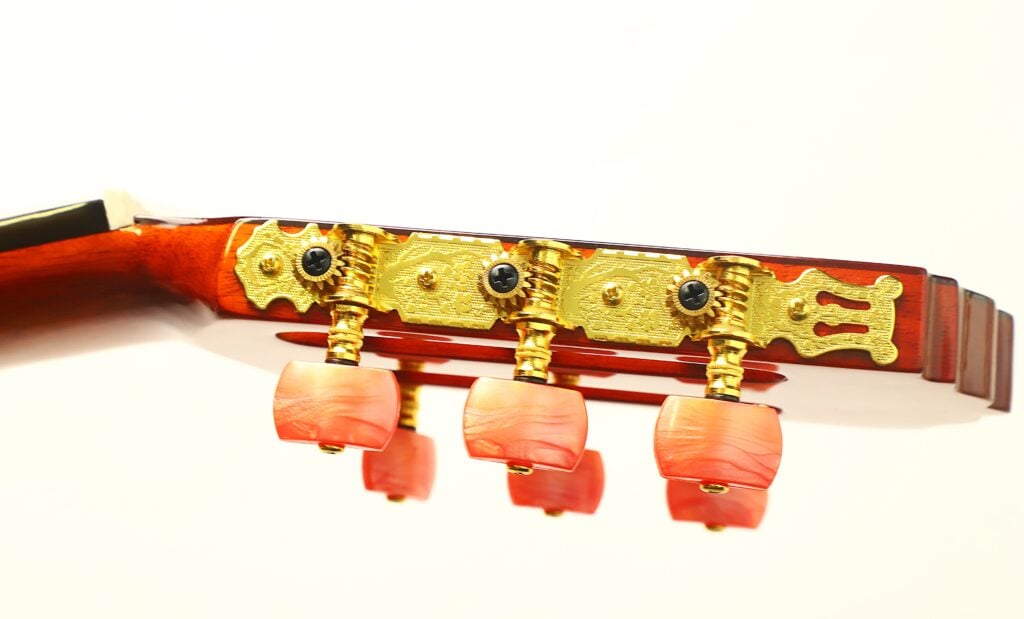 Nahaufnahme des Kopfes einer Flamenco-Gitarre von Conde Hermanos aus dem Jahr 1989 mit vergoldeten Stimmmechaniken und rot-orangefarbenen Stimmknöpfen mit Wirbelmuster. Nahaufnahme des Kopfes einer Flamenco-Gitarre von Conde Hermanos aus dem Jahr 1989 mit vergoldeten Stimmmechaniken und rot-orangefarbenen Stimmknöpfen mit Wirbelmuster.