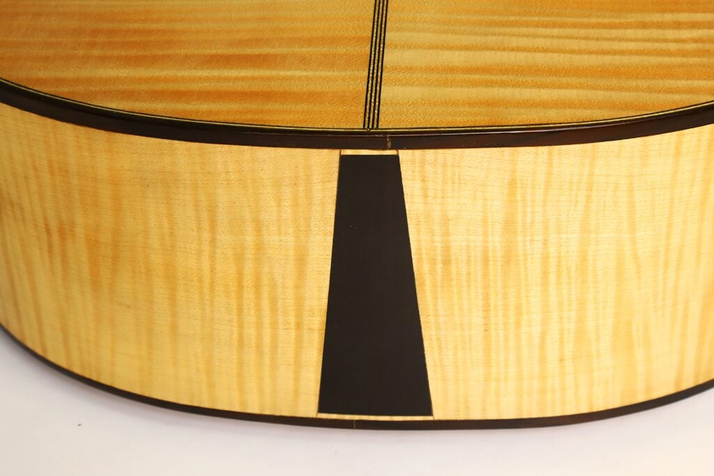 Nahaufnahme des Holzkörpers der Romantikgitarre Modell Lacote von Carlos Gass Castañeda aus dem Jahr 1995, die den dunklen Keil am Saitenhalter an der Stelle zeigt, an der die Zargen aufeinandertreffen. Nahaufnahme des Holzkörpers der Romantikgitarre Modell Lacote von Carlos Gass Castañeda aus dem Jahr 1995, die den dunklen Keil am Saitenhalter an der Stelle zeigt, an der die Zargen aufeinandertreffen.