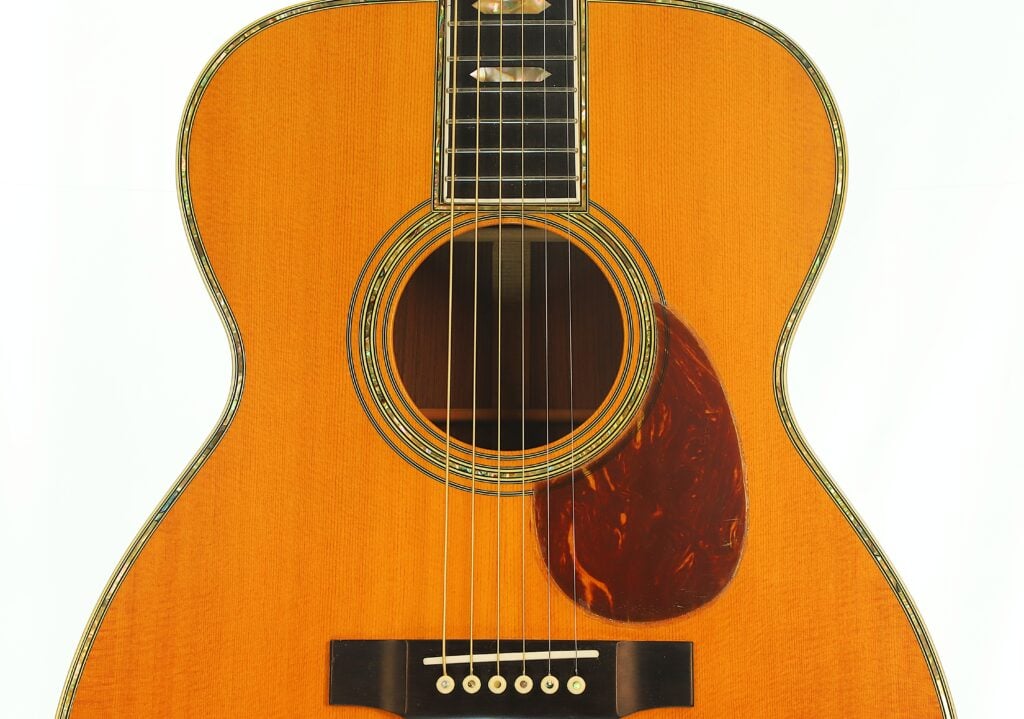 Eine Nahaufnahme des Korpus einer Martin SOM-45 Akustikgitarre aus dem Jahr 1977 mit einem dunklen Pickguard in Schildpattoptik, einem runden Schallloch mit aufwendiger Einlage und sechs Saiten, die am Steg befestigt sind. Eine Nahaufnahme des Korpus einer Martin SOM-45 Akustikgitarre aus dem Jahr 1977 mit einem dunklen Pickguard in Schildpattoptik, einem runden Schallloch mit aufwendiger Einlage und sechs Saiten, die am Steg befestigt sind.