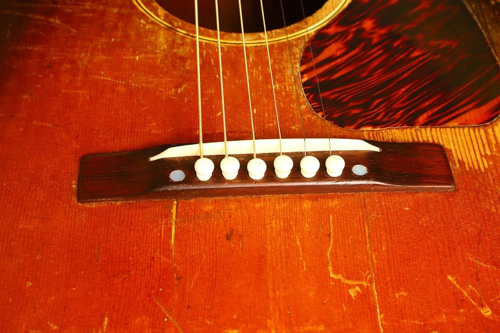Nahaufnahme des Korpus einer Gibson J-45 Banner Akustikgitarre aus dem Jahr 1943, auf der der Steg, die Saiten und ein gemustertes Schlagbrett zu sehen sind. Nahaufnahme des Korpus einer Gibson J-45 Banner Akustikgitarre aus dem Jahr 1943, auf der der Steg, die Saiten und ein gemustertes Schlagbrett zu sehen sind.