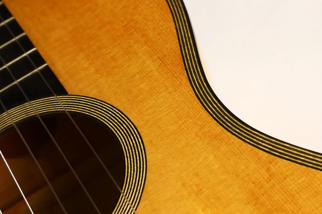 Nahaufnahme des Korpus der Modell Lacote Romantikgitarre von Carlos Gass Castañeda aus dem Jahr 1995, auf der das Schallloch, die Saiten und die gestreifte Einfassung entlang der Kante zu sehen sind. Nahaufnahme des Korpus der Modell Lacote Romantikgitarre von Carlos Gass Castañeda aus dem Jahr 1995, auf der das Schallloch, die Saiten und die gestreifte Einfassung entlang der Kante zu sehen sind.