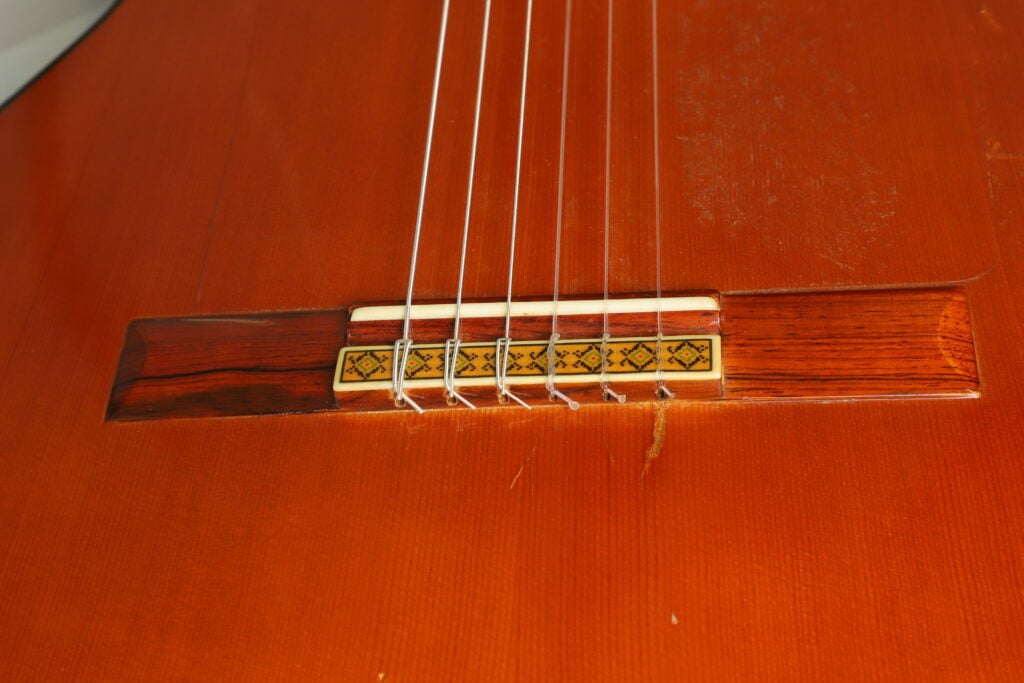 Nahaufnahme des Stegs einer Flamenco-Gitarre vom Typ Manuel Contreras 1a aus dem Jahr 1977 mit einer Zierintarsie und sechs Nylonsaiten. Nahaufnahme des Stegs einer Flamenco-Gitarre vom Typ Manuel Contreras 1a aus dem Jahr 1977 mit einer Zierintarsie und sechs Nylonsaiten.