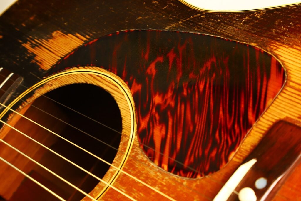 Nahaufnahme des Korpus einer Gibson J-45 Banner Akustikgitarre aus dem Jahr 1943, auf der das Schallloch, die Saiten und ein dunkel gemustertes Schlagbrett auf einer abgenutzten, strukturierten Holzoberfläche zu sehen sind. Nahaufnahme des Korpus einer Gibson J-45 Banner Akustikgitarre aus dem Jahr 1943, auf der das Schallloch, die Saiten und ein dunkel gemustertes Schlagbrett auf einer abgenutzten, strukturierten Holzoberfläche zu sehen sind.
