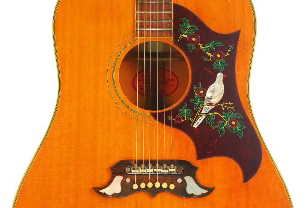 Gibson Dove 1968 Akustikgitarre mit einem dunklen Pickguard in Schildpattoptik, das mit einer Taube und einem Blumenmotiv verziert ist, sowie einem Steg mit zwei taubenförmigen Perlmutt-Einlagen. Gibson Dove 1968 Akustikgitarre mit einem dunklen Pickguard in Schildpattoptik, das mit einer Taube und einem Blumenmotiv verziert ist, sowie einem Steg mit zwei taubenförmigen Perlmutt-Einlagen.