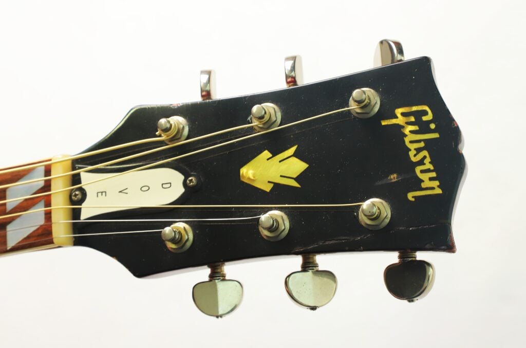 Schwarzer Kopf einer Gibson Dove Akustikgitarre aus dem Jahr 1968 mit Stimmwirbeln, einer Einlage aus Perlmutt in Form einer Taube auf der Abdeckung der Halsstange und einem goldenen Logo. Schwarzer Kopf einer Gibson Dove Akustikgitarre aus dem Jahr 1968 mit Stimmwirbeln, einer Einlage aus Perlmutt in Form einer Taube auf der Abdeckung der Halsstange und einem goldenen Logo.