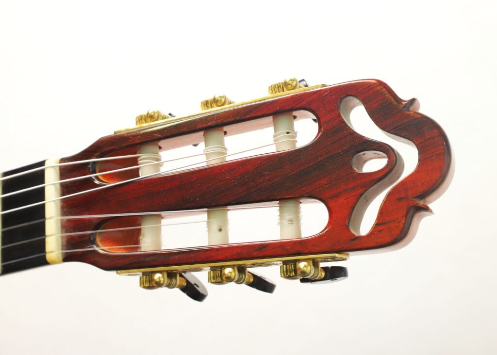 Nahaufnahme des Kopfes einer Hopf Gran Concertio 2000 Klassische Gitarre mit goldenen Stimmwirbeln, weißen Rollen und einem filigranen, geschwungenen Ausschnittmuster aus Holz. Nahaufnahme des Kopfes einer Hopf Gran Concertio 2000 Klassische Gitarre mit goldenen Stimmwirbeln, weißen Rollen und einem filigranen, geschwungenen Ausschnittmuster aus Holz.