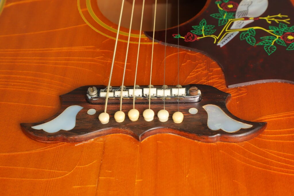 Nahaufnahme des Stegs der Gibson Dove 1968 Akustikgitarre mit Einlagen aus Perlmutt in Form einer Taube und einem Schlagbrett mit Blumen- und Vogelmotiv. Nahaufnahme des Stegs der Gibson Dove 1968 Akustikgitarre mit Einlagen aus Perlmutt in Form einer Taube und einem Schlagbrett mit Blumen- und Vogelmotiv.