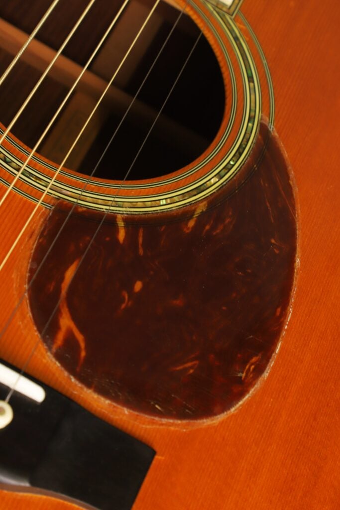 Nahaufnahme des Korpus einer Martin SOM-45 Akustikgitarre aus dem Jahr 1977, die das Schallloch, die kunstvoll verzierte Rosette und ein dunkles, gemustertes Schlagbrett im Schildpatt-Stil zeigt. Nahaufnahme des Korpus einer Martin SOM-45 Akustikgitarre aus dem Jahr 1977, die das Schallloch, die kunstvoll verzierte Rosette und ein dunkles, gemustertes Schlagbrett im Schildpatt-Stil zeigt.