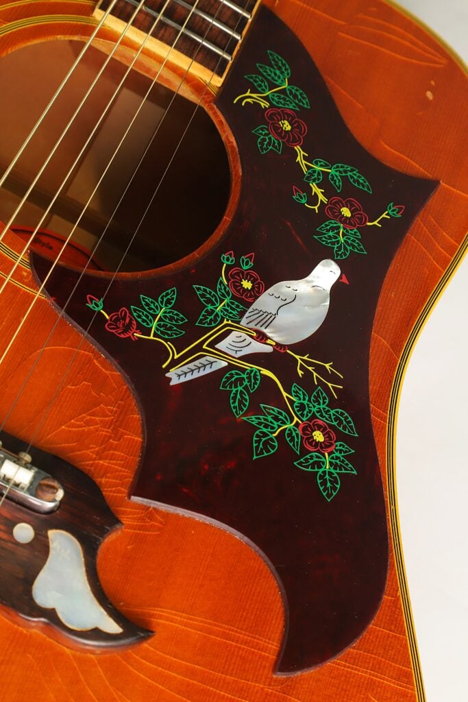 Nahaufnahme des Korpus einer Gibson Dove Akustikgitarre aus dem Jahr 1968 mit einem dunklen, schildpattfarbenen Schlagbrett, auf dem eine Taube aus Perlmutt eingelegt ist und das mit einem gemalten Blumenrankenmuster verziert ist. Nahaufnahme des Korpus einer Gibson Dove Akustikgitarre aus dem Jahr 1968 mit einem dunklen, schildpattfarbenen Schlagbrett, auf dem eine Taube aus Perlmutt eingelegt ist und das mit einem gemalten Blumenrankenmuster verziert ist.