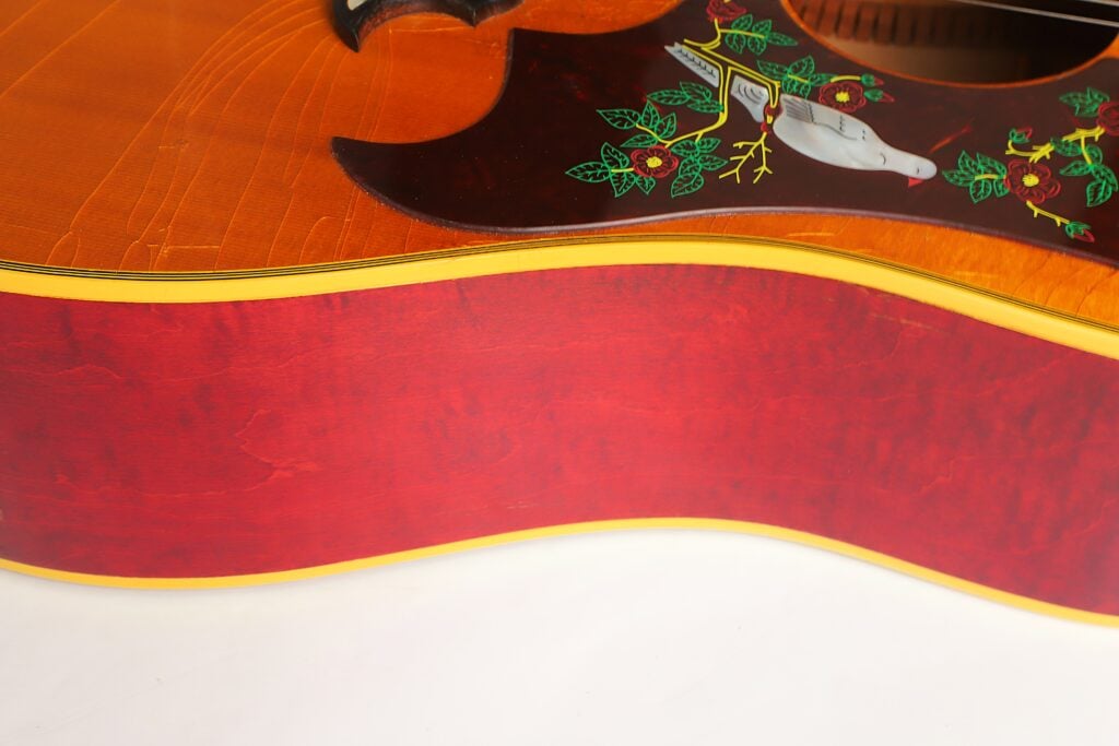 Nahaufnahme des Korpus einer Gibson Dove Akustikgitarre aus dem Jahr 1968 mit einer roten Seitenwand und einem dunklen Schlagbrett, das mit einer weißen Taube und floralen Einlegearbeiten verziert ist. Nahaufnahme des Korpus einer Gibson Dove Akustikgitarre aus dem Jahr 1968 mit einer roten Seitenwand und einem dunklen Schlagbrett, das mit einer weißen Taube und floralen Einlegearbeiten verziert ist.