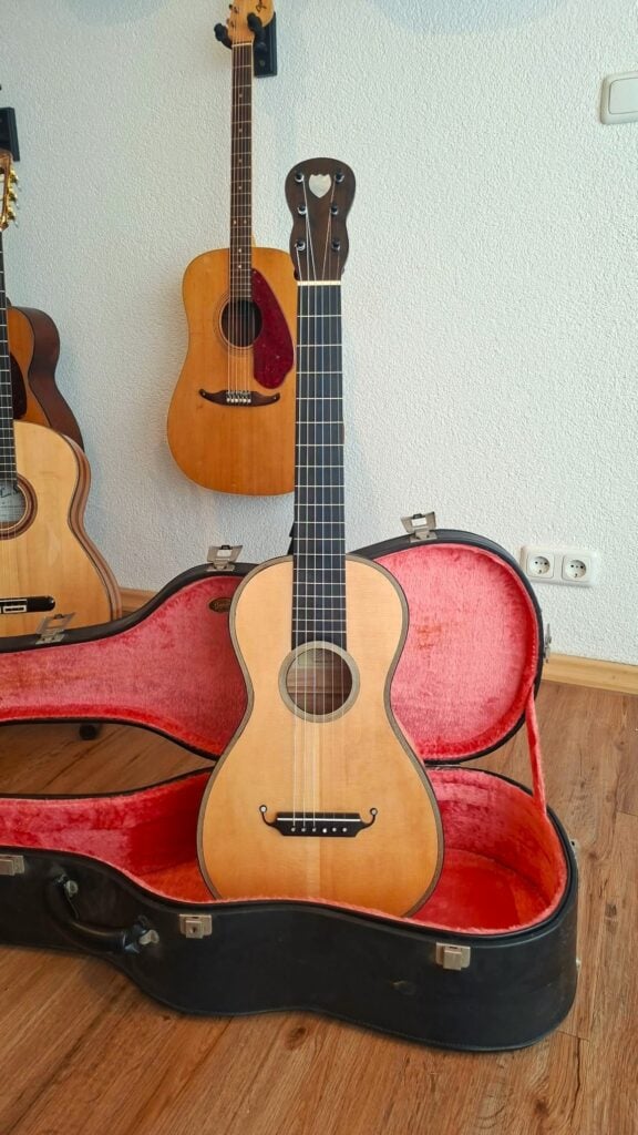 Eine Romantikgitarre des Modells Lacote von Carlos Gass Castañeda aus dem Jahr 1995 mit herzförmiger Aussparung am Kopf, die in einem offenen, rot gefütterten Koffer steht, während im Hintergrund zwei weitere Gitarren zu sehen sind. Eine Romantikgitarre des Modells Lacote von Carlos Gass Castañeda aus dem Jahr 1995 mit herzförmiger Aussparung am Kopf, die in einem offenen, rot gefütterten Koffer steht, während im Hintergrund zwei weitere Gitarren zu sehen sind.