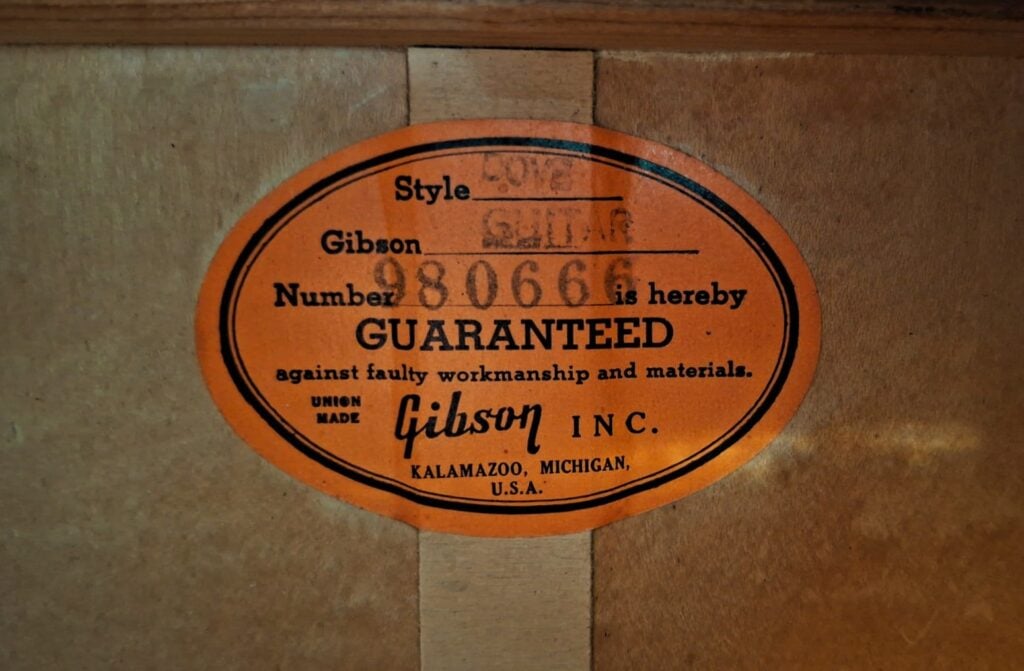Ovaler orangefarbener Aufkleber im Korpus einer Gibson Dove Akustikgitarre aus dem Jahr 1968, auf dem der Typ, die Modellnummer 980666 und Herstellerangaben vermerkt sind. Ovaler orangefarbener Aufkleber im Korpus einer Gibson Dove Akustikgitarre aus dem Jahr 1968, auf dem der Typ, die Modellnummer 980666 und Herstellerangaben vermerkt sind.