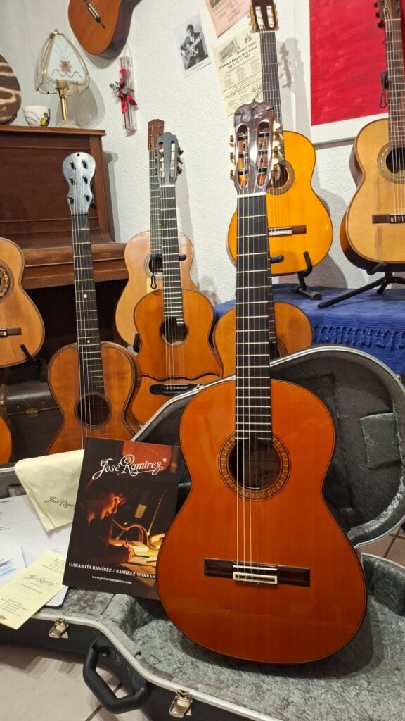 Eine Jose Ramirez 1a 2019 C-650 Tradicional Klassische Gitarre, die in ihrem offenen Hartschalenkoffer ruht, davor liegt ein Garantieschein, und im Hintergrund hängen weitere Gitarren an Wandhaltern. Eine Jose Ramirez 1a 2019 C-650 Tradicional Klassische Gitarre, die in ihrem offenen Hartschalenkoffer ruht, davor liegt ein Garantieschein, und im Hintergrund hängen weitere Gitarren an Wandhaltern.