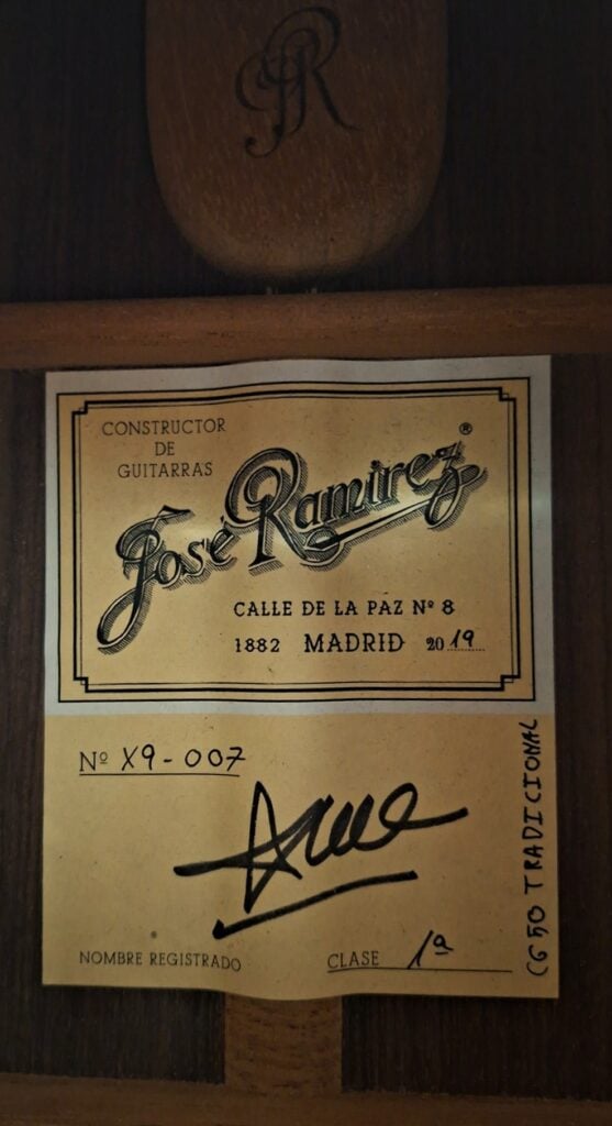 Ein gelbes, rechteckiges Etikett im Inneren einer Jose Ramirez 1a 2019 C-650 Tradicional Klassische Gitarre mit einem dekorativen Markennamen, einer Adresse in Madrid, einer handschriftlichen Seriennummer, einer Unterschrift und der Klassifizierung 1a. Ein gelbes, rechteckiges Etikett im Inneren einer Jose Ramirez 1a 2019 C-650 Tradicional Klassische Gitarre mit einem dekorativen Markennamen, einer Adresse in Madrid, einer handschriftlichen Seriennummer, einer Unterschrift und der Klassifizierung 1a.